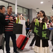 A imagem mostra uma cena em um aeroporto, possivelmente no saguão de desembarque. No centro, um grupo de pessoas está interagindo de forma alegre.

À esquerda, dois homens conversam, sorrindo. Um deles, com traços asiáticos e vestindo uma camisa preta, aponta para algo fora do quadro. O outro, com uma camisa polo listrada em vermelho, branco e azul marinho, está com uma das mãos sobre o peito, em um gesto de surpresa ou gratidão. Ambos seguram sacolas pretas com a frase "RIO GRANDE DO SUL" impressa em letras brancas.

No centro da imagem, uma mulher com um vestido de época marrom e creme, que se assemelha a trajes tradicionais gaúchos, segura duas sacolas semelhantes. Atrás dela, uma mala de rodinhas vermelha está ao lado de uma pessoa que não está completamente visível.

À direita, um artista de rua, com a pele pintada de cinza, está vestido com roupas escuras e desgastadas, incluindo um chapéu que cobre parte do seu rosto. Ele parece estar performando ou posando, e o contraste de sua figura "estática" com a alegria das outras pessoas é notável.

O fundo da imagem é o saguão do aeroporto, com janelas de vidro, portões e outras pessoas observando a cena. A iluminação é clara e uniforme, sugerindo um ambiente interno bem iluminado.