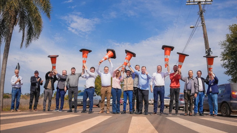 A fotografia mostra um grupo de cerca de 15 pessoas, homens e uma mulher, em pé sobre uma faixa de pedestres, celebrando uma inauguração. A maioria está segurando cones de trânsito alaranjados sobre a cabeça, como se estivessem brindando. O vice-governador Gabriel Souza está no centro da foto, com um sorriso, vestindo camisa branca de manga comprida e calça jeans escura, segurando um cone com as duas mãos. O grupo está vestido com roupas casuais. A cena ocorre em um dia ensolarado, sob um céu azul claro com nuvens. No lado direito, há um poste de energia elétrica e carros estacionados, e à esquerda, uma palmeira.