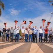 A fotografia mostra um grupo de cerca de 15 pessoas, homens e uma mulher, em pé sobre uma faixa de pedestres, celebrando uma inauguração. A maioria está segurando cones de trânsito alaranjados sobre a cabeça, como se estivessem brindando. O vice-governador Gabriel Souza está no centro da foto, com um sorriso, vestindo camisa branca de manga comprida e calça jeans escura, segurando um cone com as duas mãos. O grupo está vestido com roupas casuais. A cena ocorre em um dia ensolarado, sob um céu azul claro com nuvens. No lado direito, há um poste de energia elétrica e carros estacionados, e à esquerda, uma palmeira.
