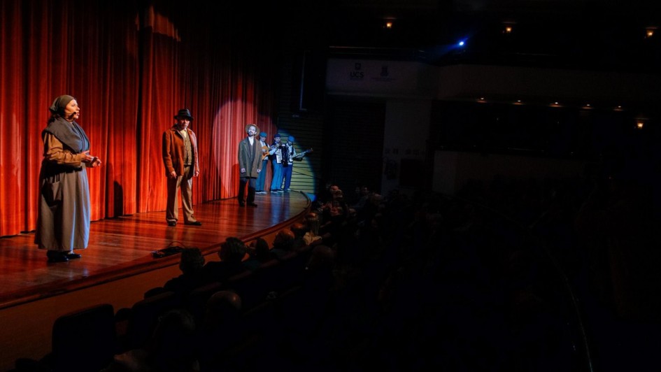 Cena de espetáculo teatral em um palco de madeira iluminado por luz amarela, com cortina vermelha ao fundo. À esquerda, uma atriz usando vestido longo cinza e lenço na cabeça fala ao público. Ao centro, dois atores trajando roupas antigas em tons terrosos estão em posição de fala, com um deles destacado por um foco de luz. Ao fundo, à direita, três músicos vestidos com boinas azuis tocam violão, sanfona e baixo. Na plateia à frente, espectadores assistem atentos em ambiente escuro.