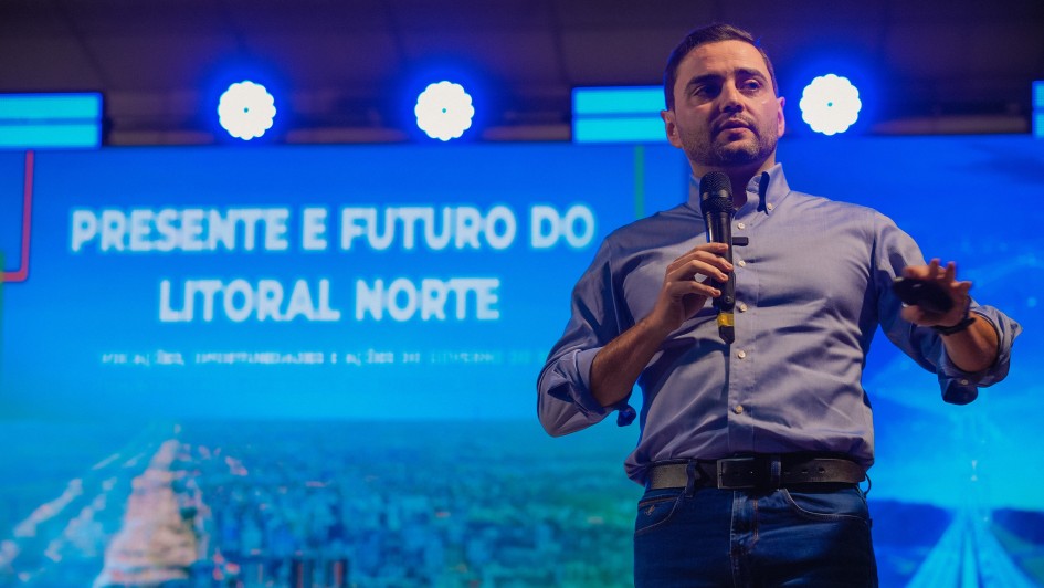 A imagem mostra o político brasileiro Gabriel Souza, vice-governador do Rio Grande do Sul, discursando em um evento. Ele está em pé, no palco, vestindo uma camisa azul clara de manga comprida e uma calça jeans escura, com um cinto preto. Ele segura um microfone com a mão direita e gesticula com a mão esquerda.

O fundo da imagem é um telão azul com a projeção de "PRESENTE E FUTURO DO LITORAL NORTE" em letras brancas e maiúsculas. Abaixo do texto, há a silhueta de uma paisagem urbana, possivelmente praias e construções do litoral, em um tom de azul mais escuro. A iluminação no palco é feita por luzes de LED brancas e redondas, criando um efeito visual que destaca o palestrante. A foto foi tirada do peito para cima, focando na figura de Gabriel Souza.