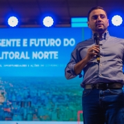A imagem mostra o político brasileiro Gabriel Souza, vice-governador do Rio Grande do Sul, discursando em um evento. Ele está em pé, no palco, vestindo uma camisa azul clara de manga comprida e uma calça jeans escura, com um cinto preto. Ele segura um microfone com a mão direita e gesticula com a mão esquerda.

O fundo da imagem é um telão azul com a projeção de "PRESENTE E FUTURO DO LITORAL NORTE" em letras brancas e maiúsculas. Abaixo do texto, há a silhueta de uma paisagem urbana, possivelmente praias e construções do litoral, em um tom de azul mais escuro. A iluminação no palco é feita por luzes de LED brancas e redondas, criando um efeito visual que destaca o palestrante. A foto foi tirada do peito para cima, focando na figura de Gabriel Souza.