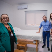 A fotografia mostra quatro pessoas, incluindo o vice-governador Gabriel Souza e a secretária da Saúde, Arita Bergmann, em uma sala de consultório médico. Da esquerda para a direita, Arita Bergmann está sorrindo, usando um blazer verde vibrante. Em seguida, Gabriel Souza, com uma camisa azul, está em pé ao lado de uma maca de exame com lençol branco. Ao seu lado, uma mulher de vestido preto e, por fim, um homem de blazer azul marinho. O ambiente é um espaço clínico novo, com paredes brancas, piso claro e uma janela com persiana.