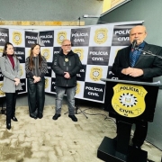 A fotografia mostra um grupo de sete pessoas, incluindo autoridades e representantes, em pé, posando para uma foto de grupo em uma inauguração. Um homem careca, com barba e terno escuro, está no lado direito, falando em um púlpito com a insígnia da Polícia Civil. O grupo atrás dele é composto por um oficial militar com boina, uma mulher de blazer branco, um homem de terno cinza, e duas mulheres com casacos escuros. Eles estão em frente a um banner com o logo e o nome da "Polícia Civil" repetidamente. 