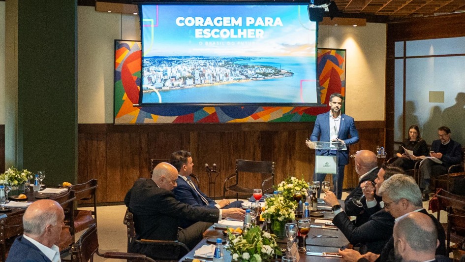 A imagem retrata uma palestra com diversas pessoas sentadas ao longo de mesas retangulares, todas em trajes sociais. Na frente do ambiente, uma tela grande exibe a frase "Coragem para escolher" sobre uma imagem de paisagem urbana, enquanto o governador apresenta no púlpito.