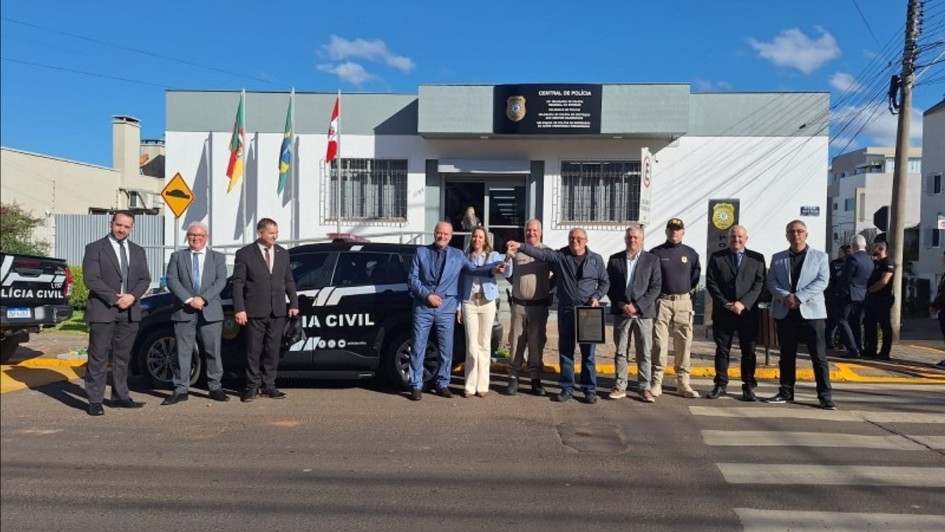 A foto mostra um grupo de cerca de dez pessoas posando em frente a um prédio da Polícia Civil em Vacaria. Eles estão em pé, com uma viatura policial preta visível à esquerda, e um prédio branco e cinza com a inscrição "CENTRAL DE POLÍCIA" e o logo da "POLÍCIA CIVIL" acima da porta. No centro da imagem, dois homens seguram um quadro. À frente do grupo, há uma faixa de pedestres. As bandeiras do Brasil e do Rio Grande do Sul estão hasteadas na lateral da delegacia. O dia está ensolarado com céu azul e algumas nuvens.