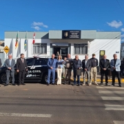 A foto mostra um grupo de cerca de dez pessoas posando em frente a um prédio da Polícia Civil em Vacaria. Eles estão em pé, com uma viatura policial preta visível à esquerda, e um prédio branco e cinza com a inscrição "CENTRAL DE POLÍCIA" e o logo da "POLÍCIA CIVIL" acima da porta. No centro da imagem, dois homens seguram um quadro. À frente do grupo, há uma faixa de pedestres. As bandeiras do Brasil e do Rio Grande do Sul estão hasteadas na lateral da delegacia. O dia está ensolarado com céu azul e algumas nuvens.