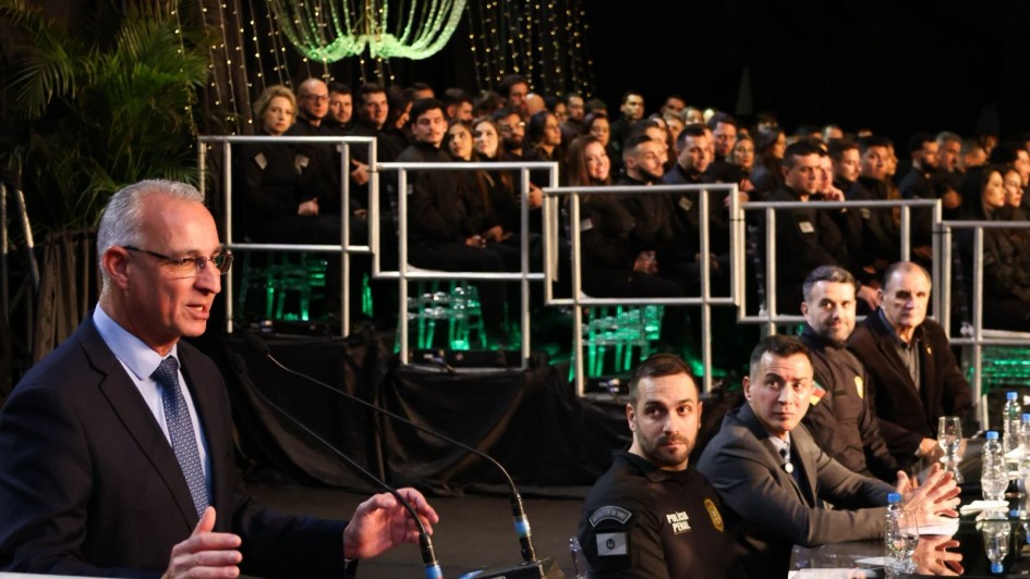 A imagem retrata uma formatura em um auditório, com várias pessoas sentadas em plataformas elevadas ao fundo, decoradas com iluminação verde e elementos suspensos. Em primeiro plano, um homem de terno fala ao microfone em um púlpito, enquanto à direita da imagem estão pessoas em trajes formais e uniformes policiais, sentadas à mesa.