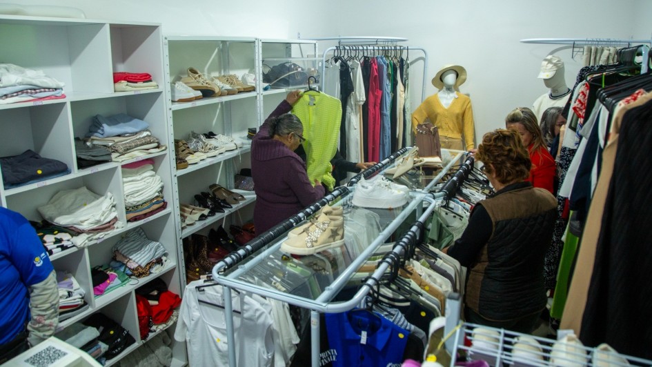 A fotografia retrata o interior de um bazar social. O espaço está repleto de prateleiras e araras com roupas, sapatos e outros itens, organizados para venda. Duas mulheres estão em pé, olhando as roupas em uma arara no centro. Uma delas, de cabelo curto, segura uma blusa verde-limão. Ao fundo, manequins exibem algumas roupas e um ventilador de teto é visível no teto.