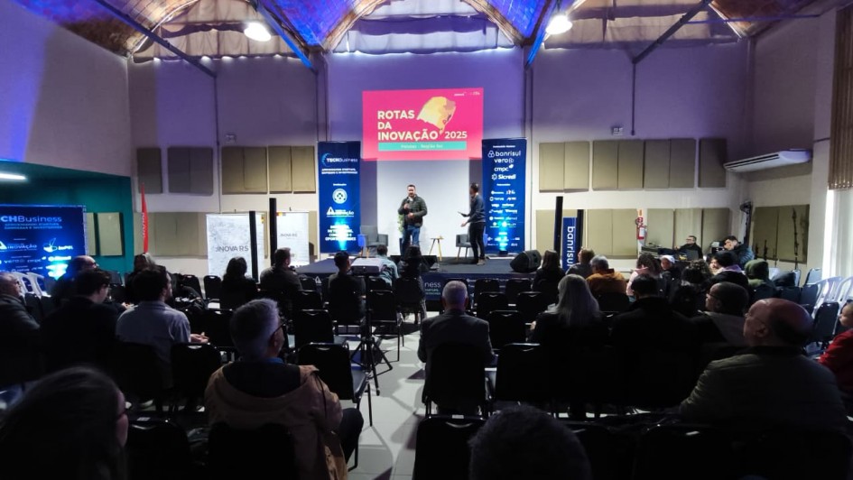 Foto tirada a partir dos fundos de um auditório, focando o palco do evento. Na metade inferior do quadro, estão várias pessoas sentadas. Ao fundo, um homem fala no palco. Atrás dele há um painel no qual está escrito: "Rotas da Inovação 2025".