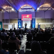 Foto tirada a partir dos fundos de um auditório, focando o palco do evento. Na metade inferior do quadro, estão várias pessoas sentadas. Ao fundo, um homem fala no palco. Atrás dele há um painel no qual está escrito: "Rotas da Inovação 2025".