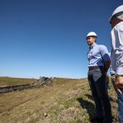 Dois homens usam capacete de proteção branco e roupas sociais casuais, observando a estrutura de uma barragem em área rural. Um deles está de camisa azul clara e calça cinza, enquanto o outro veste camisa branca e calça jeans. O céu está limpo e azul, e a cena transmite a ideia de vistoria técnica ou acompanhamento de obra. Ao fundo, é possível ver parte da estrutura de concreto da barragem e áreas de vegetação.