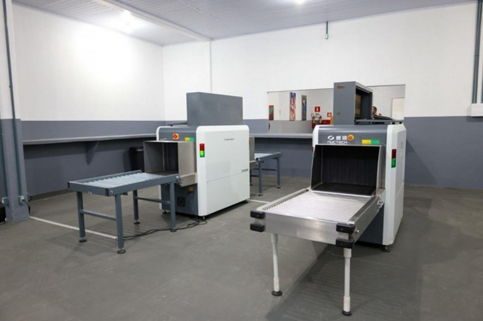 A foto mostra o interior de um prédio, que aparenta ser uma sala de revista em um complexo prisional, com dois equipamentos de raio-x para a inspeção de objetos. As máquinas são brancas com detalhes em cinza e preto, e possuem esteiras para o transporte dos itens. A sala tem paredes brancas e um chão cinza. Ao fundo, uma bancada cinza se estende ao longo da parede, e um espelho reflete uma parte do ambiente. A sala é limpa, organizada e bem iluminada por luzes de teto brancas.