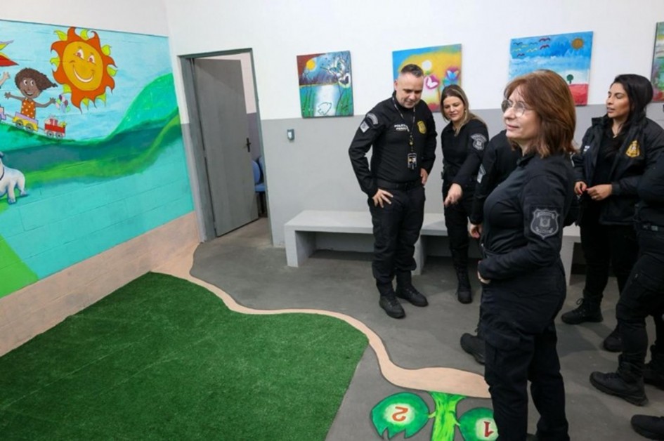 A foto mostra um grupo de cinco pessoas em uma sala com decoração infantil, aparentemente um espaço de visita para crianças. O ambiente tem paredes decoradas com pinturas de paisagens e personagens de desenhos animados. No chão, há um tapete de grama sintética verde com um caminho de terra de mentira, e as imagens de árvores e números. As pessoas na foto estão em pé, vestindo uniformes pretos, observando e conversando. No fundo, há um banco branco de concreto e uma porta cinza. A iluminação é clara e a cena sugere uma vistoria no local.