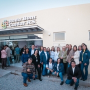 Um grupo de pessoas posa para foto em frente a um prédio de fachada clara, identificado com a placa “Rede Bem Cuidar RS”. A entrada é de vidro, e atrás dela aparecem outras pessoas no interior do espaço. Em destaque, cerca de 20 pessoas estão reunidas do lado de fora, sorrindo para a câmera. Algumas usam jalecos brancos e camisetas do programa, enquanto outras estão de casaco ou roupas sociais. No canto inferior esquerdo, um homem agachado faz sinal de positivo. O cenário é de inauguração ou evento oficial, em ambiente externo com piso de brita e calçamento.
