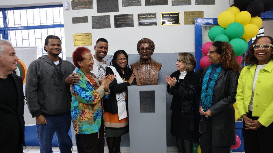 Diversas pessoas estão reunidas em torno do busto recém-inaugurado do poeta Oliveira Silveira. Elas batem palmas e sorriem para a foto. 