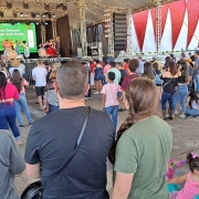 Foto mostra o público de costa, várias pessoas olhando para um palco ao fundo, onde acontece uma apresentação de dança tradicionalista gaúcha. 