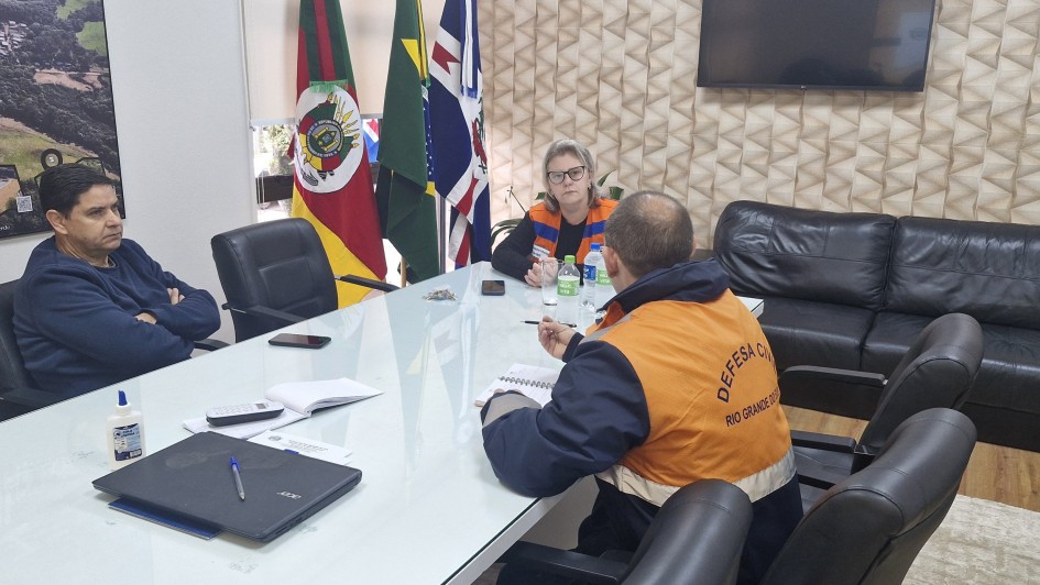 Três pessoas estão reunidas ao redor de uma mesa retangular em uma sala. Duas delas vestem coletes da Defesa Civil.