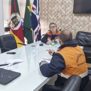 Três pessoas estão reunidas ao redor de uma mesa retangular em uma sala. Duas delas vestem coletes da Defesa Civil. 