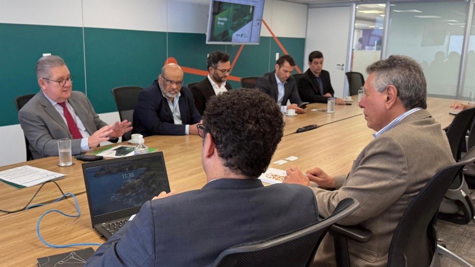 A imagem mostra uma reunião corporativa em uma sala de conferência moderna. Nove homens de terno estão sentados ao redor de uma mesa retangular de madeira clara. No canto esquerdo da imagem, um homem de cabelos grisalhos e óculos, vestido com terno cinza e gravata vermelha, está falando enquanto gesticula com as mãos. À sua frente há um copo com água e alguns papéis.
No centro da mesa, outro homem com cabelo escuro e encaracolado está de costas para a câmera, trabalhando em um notebook que exibe a hora "11:36". Vários outros participantes estão atentos ou usando seus dispositivos eletrônicos, como celulares e laptops. Ao fundo, uma televisão está presa à parede exibindo uma apresentação com o título “Brasil” e o logo da Finep (Financiadora de Estudos e Projetos), sugerindo que a reunião seja relacionada a negócios ou projetos de inovação e desenvolvimento no Brasil.
A sala é bem iluminada, com paredes brancas e divisórias de vidro, transmitindo um ambiente profissional e organizado.