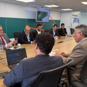 A imagem mostra uma reunião corporativa em uma sala de conferência moderna. Nove homens de terno estão sentados ao redor de uma mesa retangular de madeira clara. No canto esquerdo da imagem, um homem de cabelos grisalhos e óculos, vestido com terno cinza e gravata vermelha, está falando enquanto gesticula com as mãos. À sua frente há um copo com água e alguns papéis.

No centro da mesa, outro homem com cabelo escuro e encaracolado está de costas para a câmera, trabalhando em um notebook que exibe a hora "11:36". Vários outros participantes estão atentos ou usando seus dispositivos eletrônicos, como celulares e laptops. Ao fundo, uma televisão está presa à parede exibindo uma apresentação com o título “Brasil” e o logo da Finep (Financiadora de Estudos e Projetos), sugerindo que a reunião seja relacionada a negócios ou projetos de inovação e desenvolvimento no Brasil.

A sala é bem iluminada, com paredes brancas e divisórias de vidro, transmitindo um ambiente profissional e organizado.