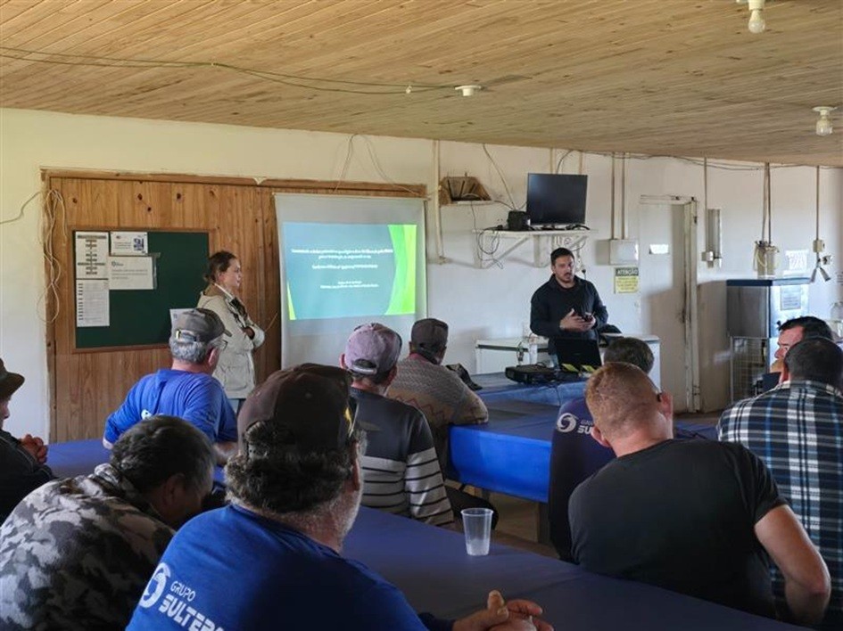 Foto em ambiente interno. Há diversos homens sentados de frente para um homem que apresenta uma palestra. No fundo, há uma tela projetada com slides.
