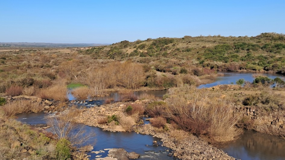 Foto do arroio Taquarembó em um dia de céu azul. As margens estão com vegetações rasteiras de cor marrom e verde.
