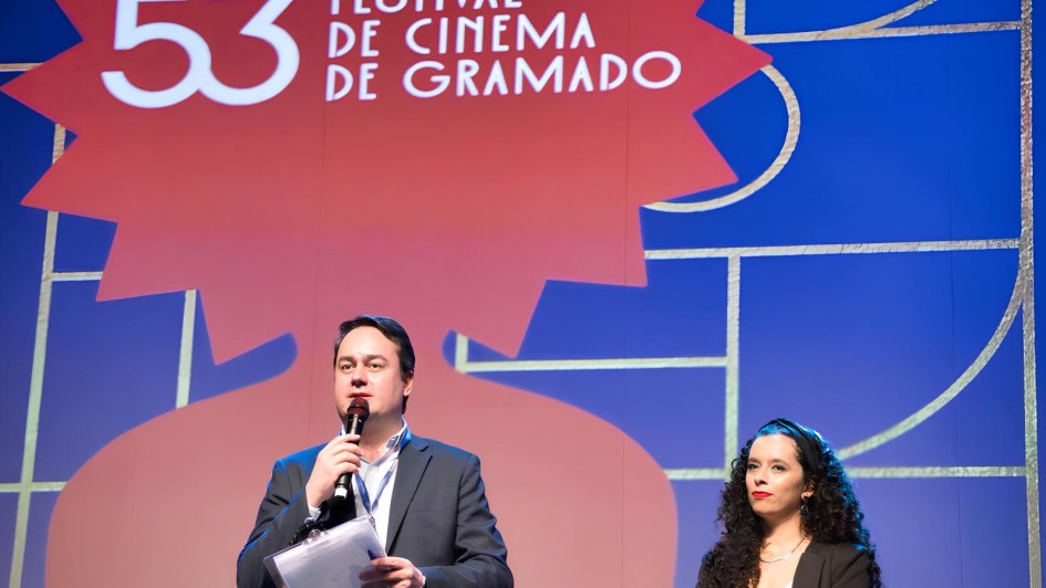 Fotografia tirada durante o 53º Festival de Cinema de Gramado. A imagem mostra um homem e uma mulher no palco do evento. O homem está à esquerda, segurando um microfone com uma das mãos e alguns papéis com a outra. Ele veste um terno escuro com camisa clara e fala ao microfone. A mulher está à direita, com cabelos cacheados soltos, longos e escuros. Ela veste um blazer preto sobre uma blusa clara e está olhando em direção ao público, com expressão séria.
Ao fundo, há um grande painel colorido em tons de azul e vermelho, com o logo do festival no topo: "53 Festival de Cinema de Gramado". O design gráfico ao fundo remete ao tradicional troféu Kikito, símbolo do evento. A iluminação é intensa, destacando os apresentadores e o logotipo do festival.