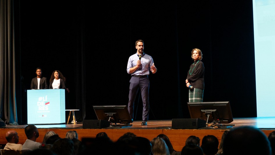 Evento com público: um homem de camisa azul claro fala ao microfone no centro de um palco, ao lado de uma mulher de saia xadrez. À esquerda, dois outros participantes estão atrás de um púlpito azul com a hashtag "#LÍDERES". A plateia está sentada, observando. O ambiente é escuro, com telas iluminadas ao fundo.
