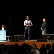 Evento com público: um homem de camisa azul claro fala ao microfone no centro de um palco, ao lado de uma mulher de saia xadrez. À esquerda, dois outros participantes estão atrás de um púlpito azul com a hashtag "#LÍDERES". A plateia está sentada, observando. O ambiente é escuro, com telas iluminadas ao fundo.