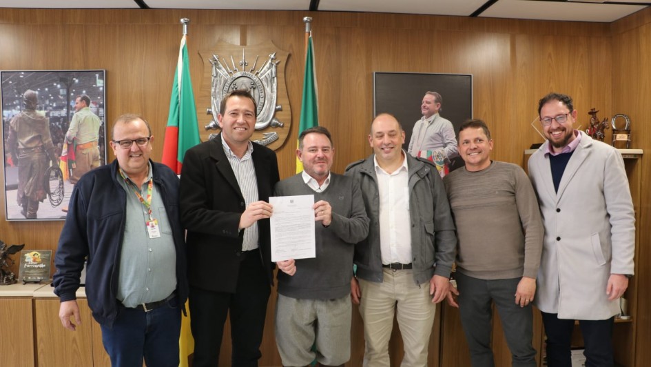 Seis homens posam para a foto, lado a lado, sorrindo, em uma sala de reuniões. Ao centro, dois deles seguram um documento.