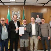 Seis homens posam para a foto, lado a lado, sorrindo, em uma sala de reuniões. Ao centro, dois deles seguram um documento. 