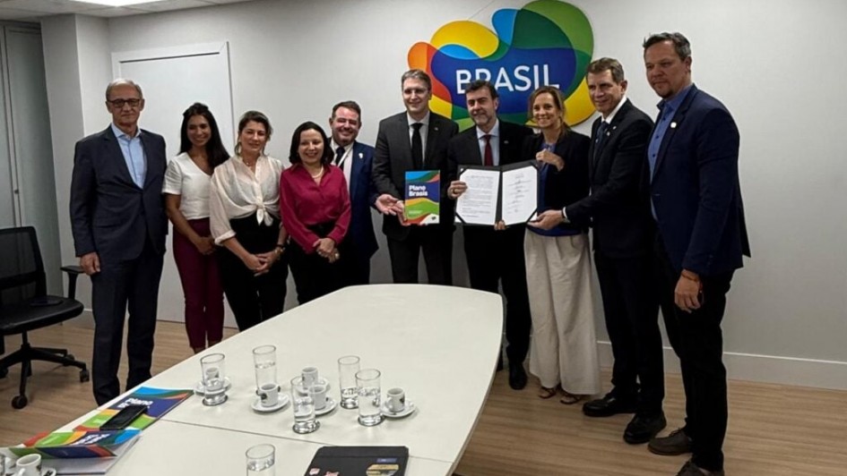 Foto em ambiente fechado de dez pessoas posando de frente, em pé. As que estão ao meio seguram o documento assinado, exibindo-o. Na parte inferior da foto, está uma mesa branca com copos.
