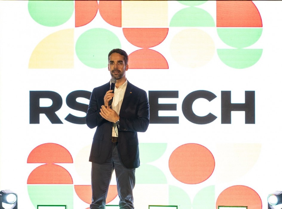 O governador Eduardo Leite está ao centro da imagem, em pé, falando ao microfone. Ao fundo, há um painel iluminado e colorido (nas cores verde, amarelo e vermelho), com a identidade visual do RS Tech.
