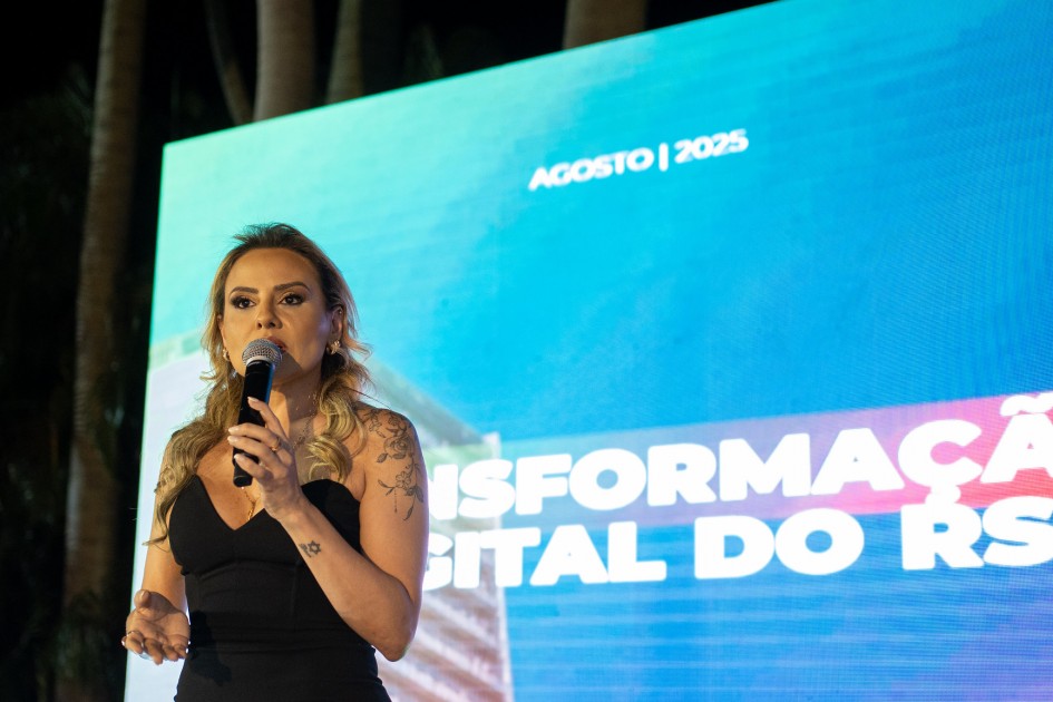 A titular da SPGG, Danielle Calazans, fala ao microfone em frente a um painel iluminado e colorido.