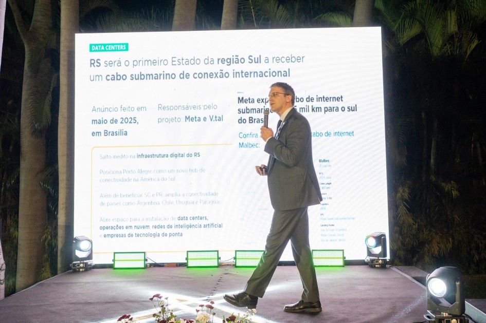 Um homem de terno fala ao microfone em frente a um painel iluminado que exibe uma apresentação sobre data centers.