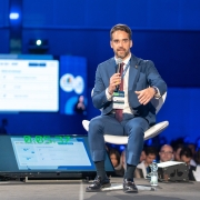 Homem de terno azul e gravata vinho está sentado em uma cadeira branca, falando ao microfone em um palco de evento. Ele segura o microfone com uma das mãos e faz gesto com a outra. Usa crachá de identificação pendurado no pescoço. À frente dele, há um monitor digital exibindo informações e uma contagem regressiva em verde. No fundo, um telão desfocado projeta dados e logotipos. O ambiente está iluminado em tons de azul e o público aparece desfocado na plateia.