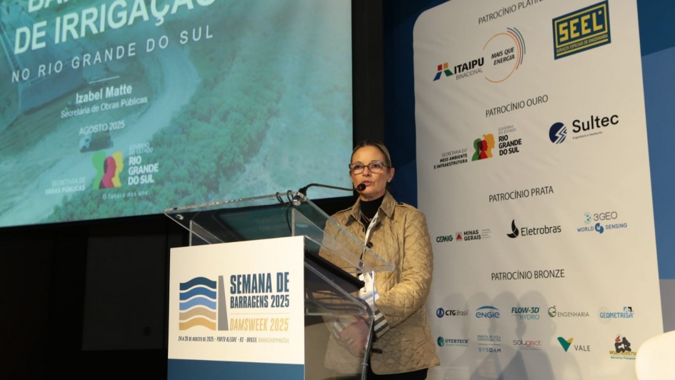 Foto de Izabel falando ao púlpito, em um palco. Atrás dela, há um painel com a logo dos órgãos envolvidos no evento. Do outro lado, também ao fundo, há uma apresentação de slide com os dizeres: "Barragens de irrigação no Rio Grande do Sul".