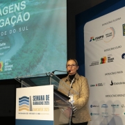 Foto de Izabel falando ao púlpito, em um palco. Atrás dela, há um painel com a logo dos órgãos envolvidos no evento. Do outro lado, também ao fundo, há uma apresentação de slide com os dizeres: "Barragens de irrigação no Rio Grande do Sul".
