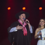 Um homem e uma mulher cantam no palco durante um show. O homem veste traje tradicional gaúcho, incluindo chapéu preto, poncho escuro, camisa listrada e lenço vermelho no pescoço. Ele segura um microfone e canta com expressão séria. Ao lado, a mulher usa vestido branco de um ombro só, com cinto dourado, e também segura um microfone, sorrindo enquanto canta. Ao fundo, luzes laranjas iluminam o cenário.