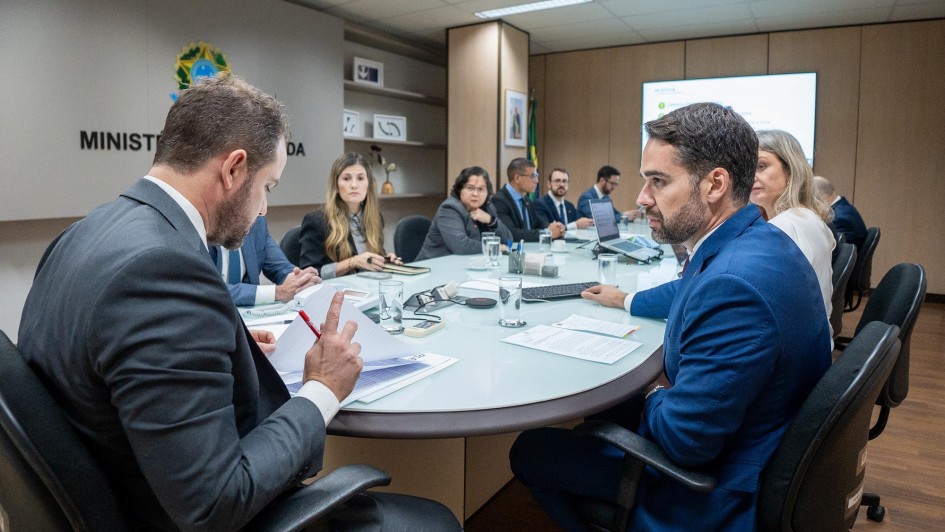 Reunião em uma sala do Ministério da Fazenda. Várias pessoas estão sentadas ao redor de uma mesa redonda, com computadores, documentos e copos de água à frente. Em destaque, dois homens em primeiro plano, ambos de terno, um deles segurando papéis e caneta, enquanto o outro observa atentamente. Ao fundo, outras pessoas acompanham a discussão, e um telão exibe informações projetadas. Na parede, aparece parcialmente o brasão da República e a inscrição “Ministério da Fazenda”.