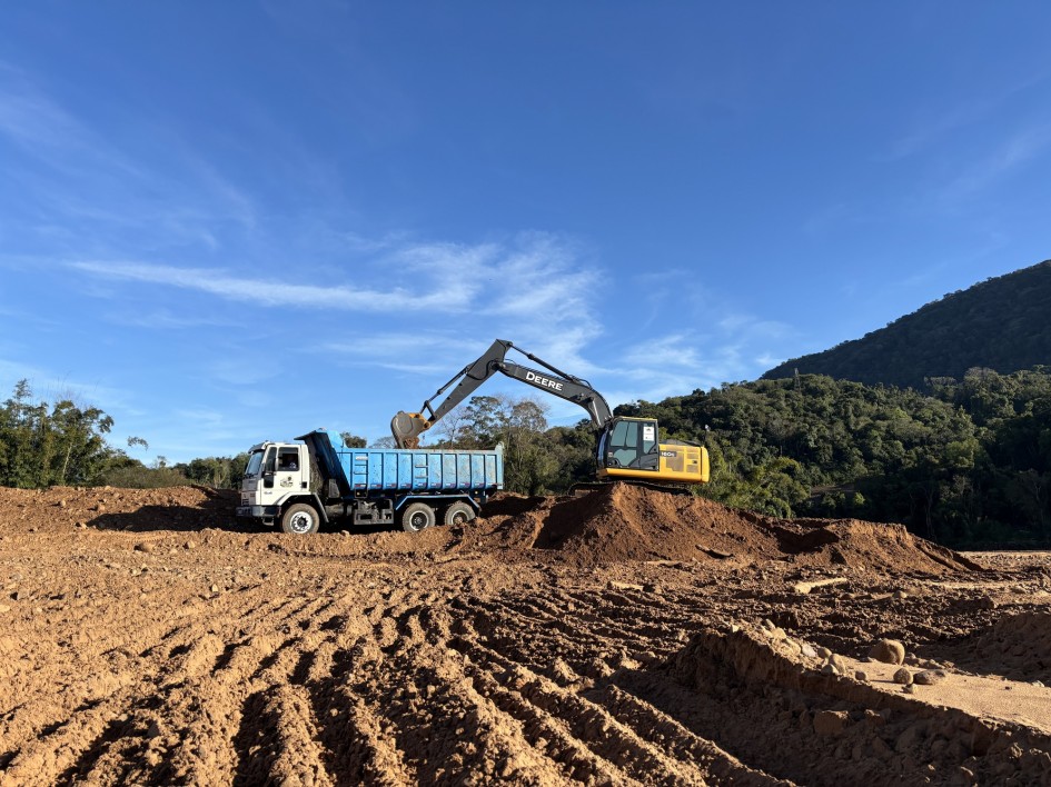 A imagem mostra um cenário de terra batida sob um céu azul. Um caminhão basculante branco e azul, com a caçamba erguida, está sendo carregado por uma escavadeira amarela da marca John Deere. A escavadeira está no topo de uma pilha de terra. Ao fundo, há uma área de mata fechada e um morro coberto de vegetação. A luz do sol incide sobre a cena, projetando algumas sombras. A terra, de cor marrom, apresenta marcas de pneus e tratores.