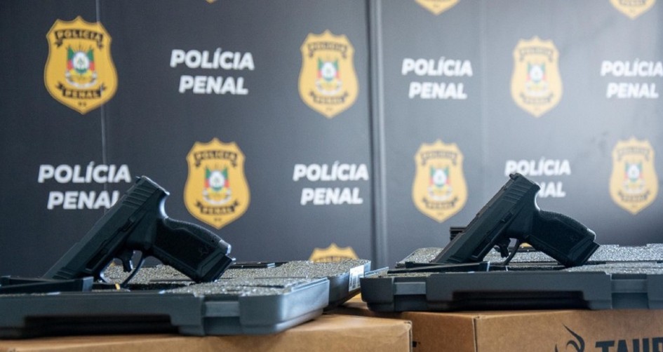 Foto mostra duas pistolas pretas em primeiro plano. Ao fundo, uma parede preta com várias logos da polícia penal, num escudo amarelo, com o brasão do Rio Grande do Sul, no meio.
