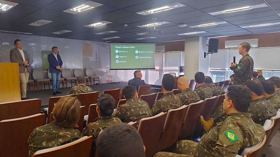 Em um auditório, dezenas de militares do Exército Brasileiro, com fardas camufladas e o brasão com a bandeira do Brasil na manga, estão sentados de costas para a câmera. À direita, um militar em pé fala ao microfone. À esquerda do palco, dois homens de terno acompanham a fala ao lado de um púlpito e de cadeiras vazias. Ao fundo, um telão projeta um slide verde com o texto “Sigam nossas redes” e ícones de redes sociais; o ambiente é iluminado por luzes de teto e por janelas laterais.