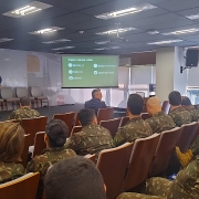 Em um auditório, dezenas de militares do Exército Brasileiro, com fardas camufladas e o brasão com a bandeira do Brasil na manga, estão sentados de costas para a câmera. À direita, um militar em pé fala ao microfone. À esquerda do palco, dois homens de terno acompanham a fala ao lado de um púlpito e de cadeiras vazias. Ao fundo, um telão projeta um slide verde com o texto “Sigam nossas redes” e ícones de redes sociais; o ambiente é iluminado por luzes de teto e por janelas laterais.