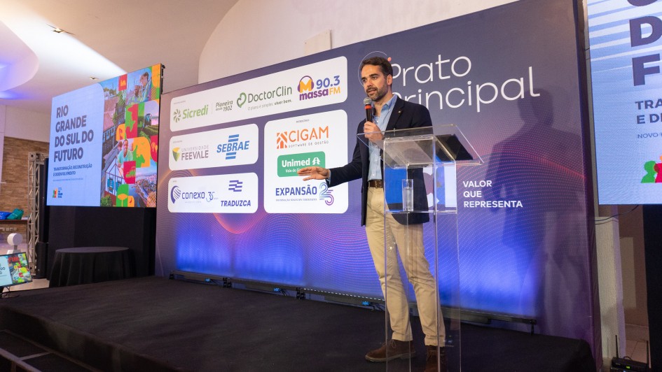 A imagem retrata uma apresentação em um palco, onde o governador está em pé, segurando um microfone e falando para a plateia. Ao fundo, há um grande painel com logotipos de diversas empresas e instituições. À esquerda, uma tela de projeção exibe o título "Rio Grande do Sul do Futuro" acompanhado de gráficos coloridos.