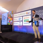 A imagem retrata uma apresentação em um palco, onde o governador está em pé, segurando um microfone e falando para a plateia. Ao fundo, há um grande painel com logotipos de diversas empresas e instituições. À esquerda, uma tela de projeção exibe o título "Rio Grande do Sul do Futuro" acompanhado de gráficos coloridos.