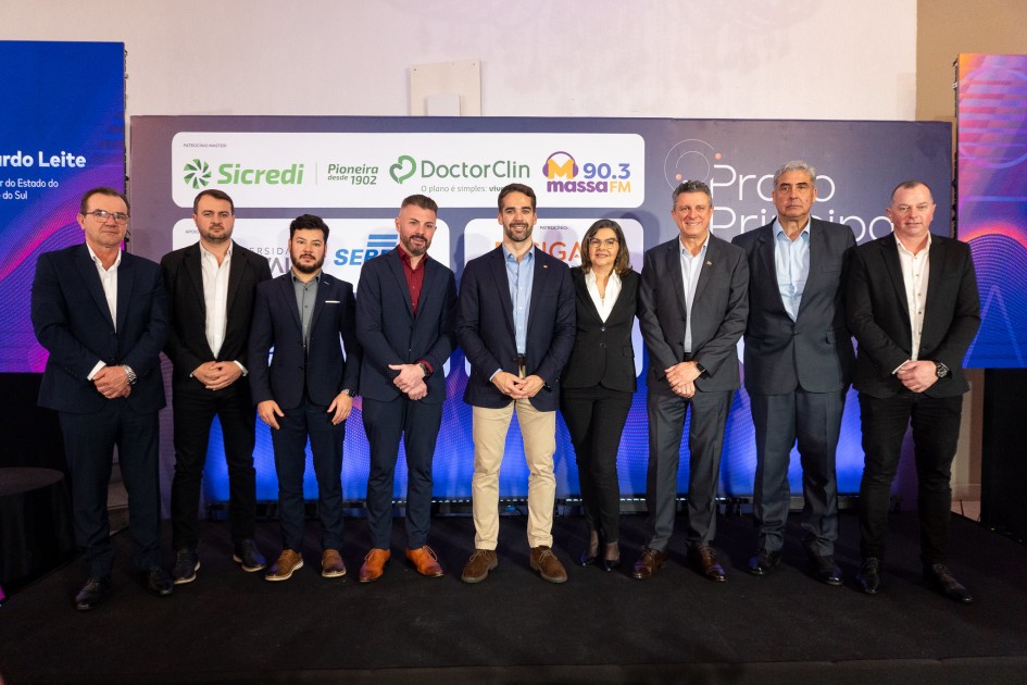A imagem mostra nove pessoas em pé, lado a lado, posando para uma foto em frente a um painel com logotipos de diversas empresas e instituições. O governador está no centro da imagem. Todos estão vestidos com trajes formais.