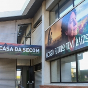 Fachada de prédio com o nome “Casa da Secom”. Um grande banner mostra cenas rurais com galinhas, pessoas no campo e o rosto de uma mulher, acompanhado da frase: “Nosso futuro tem raízes fortes”. O ambiente transmite conexão com o campo e comunicação pública.