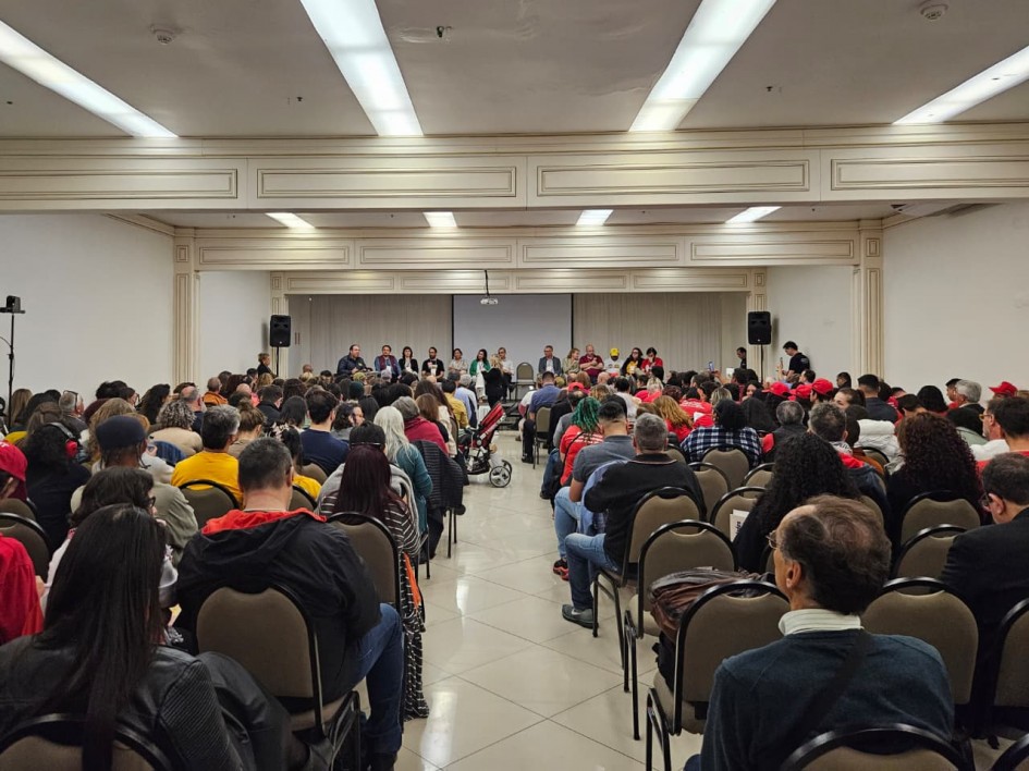 Imagem de um auditório lotado. Há diversos homens e mulheres sentados no palco, de frente para uma plateia lotada, que aparece de costas na imagem.